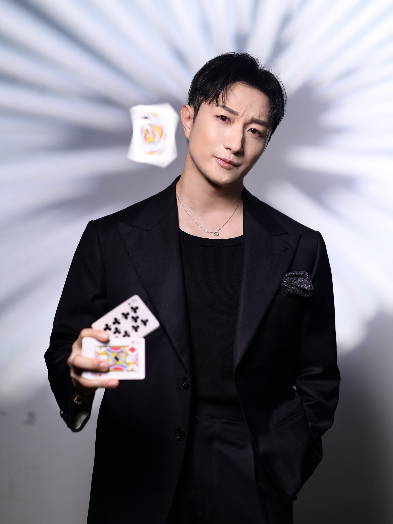 John Fung in a black suit holding two cards as a third spins in mid-air, light streaking behind him